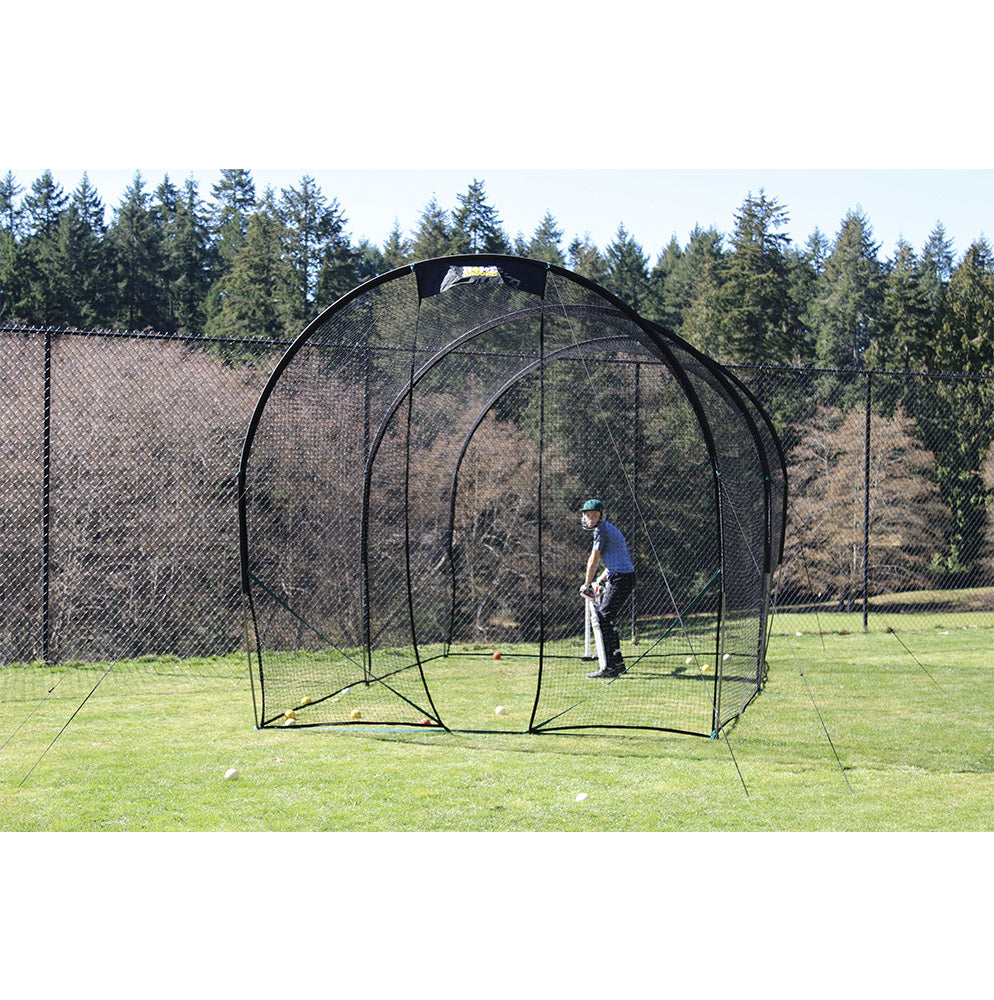 A person is practicing cricket batting inside a black colored portable cricket net set up on a grassy field.