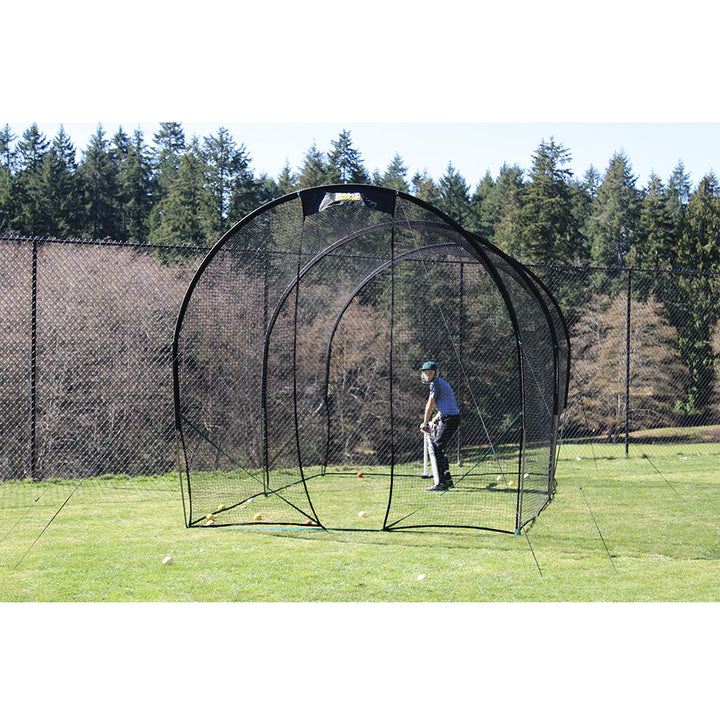 A person is practicing cricket batting inside a black colored portable cricket net set up on a grassy field.