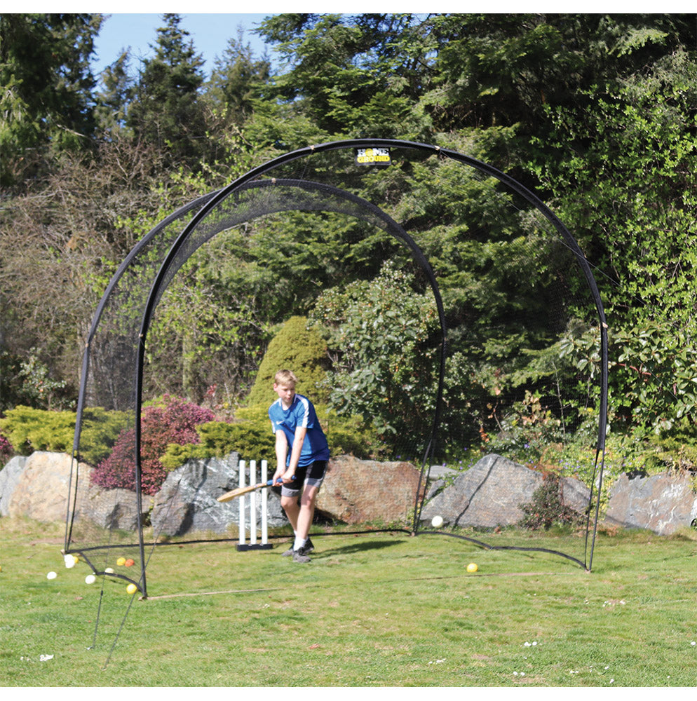 A person is practicing cricket batting against a cricket net which is set up outdoors. There are cricket balls visible in the air and on the ground.