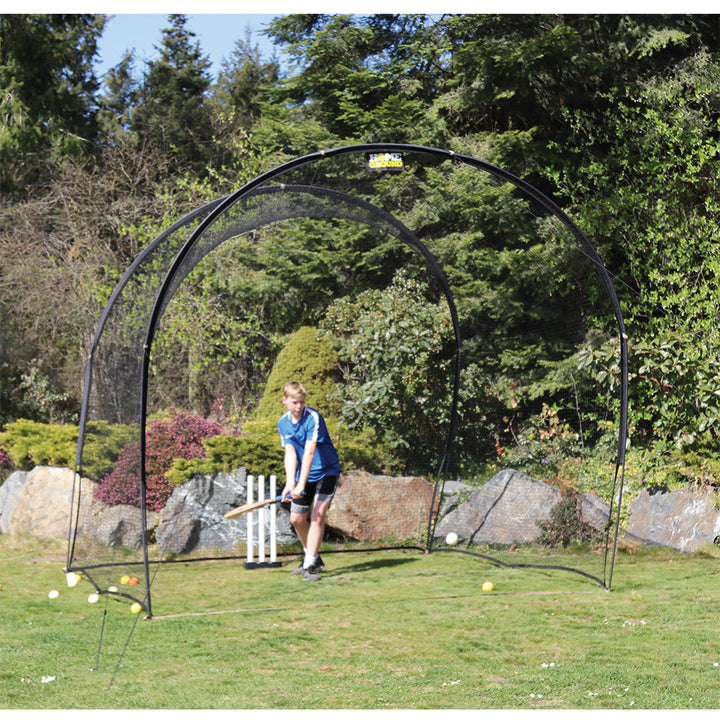A person is practicing cricket batting against a cricket net which is set up outdoors. There are cricket balls visible in the air and on the ground.