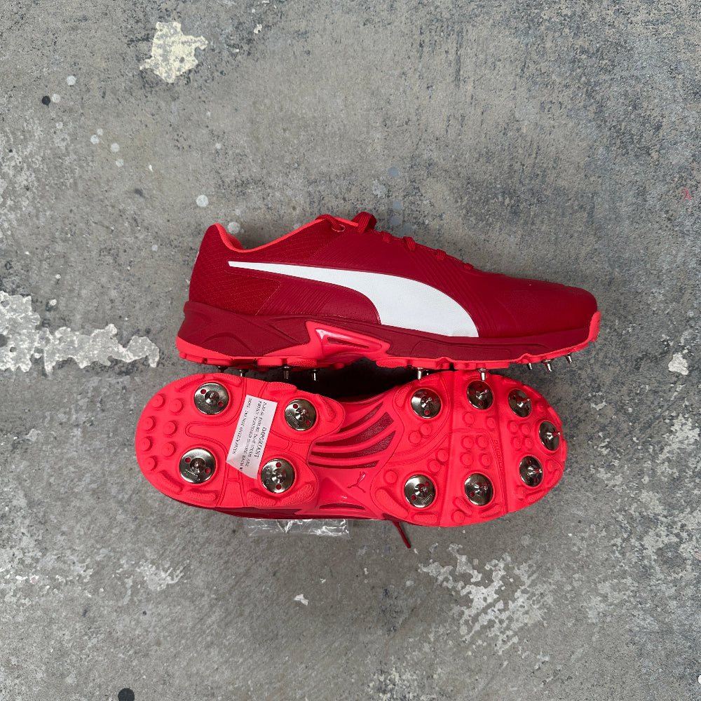 Red Puma cricket shoe with spikes on a gray concrete surface