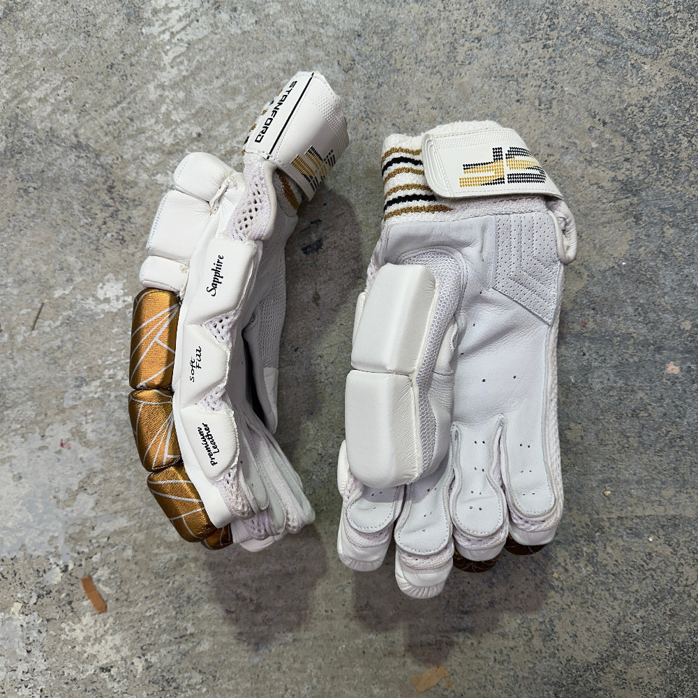 Pair of white SF cricket gloves with gold accents on a gray concrete surface