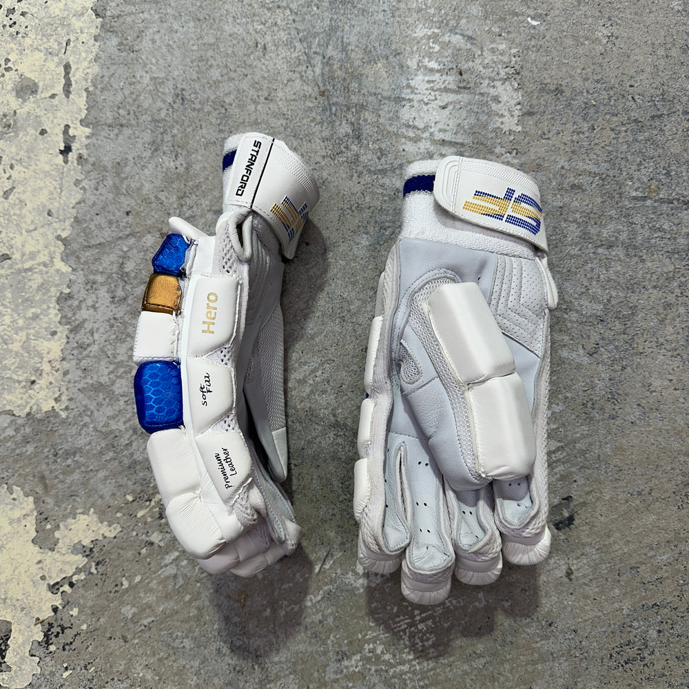 Pair of white SF cricket gloves with blue and gold accents on a gray concrete surface