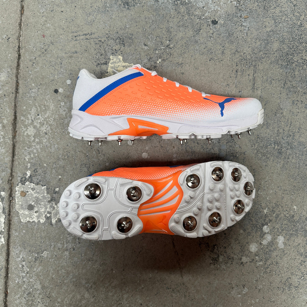 Orange and white Puma cricket shoes with spikes on a concrete surface