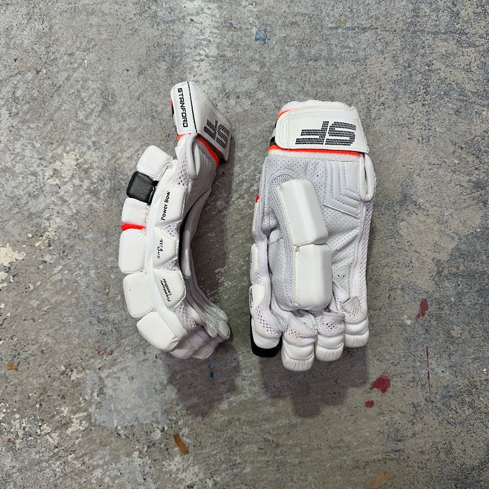 Pair of white cricket batting gloves with black and orange accents on a gray concrete surface with SF branding.