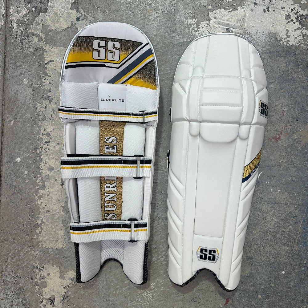 Pair of white SS cricket pads with gold and black accents on a gray concrete surface