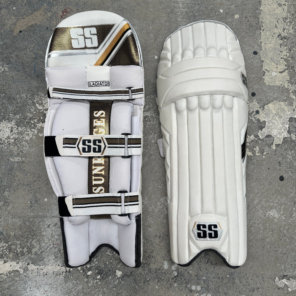 White cricket pads with black and gold SS branding on a textured gray background