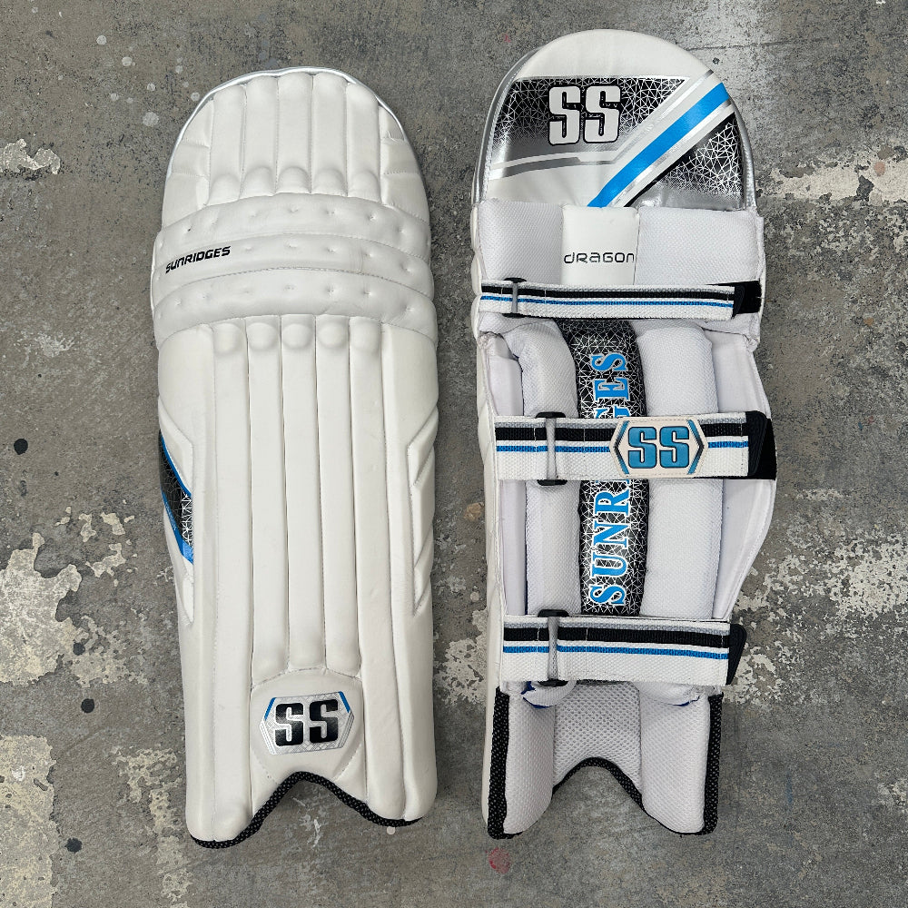 Pair of white SS Dragon cricket pads with branding on a textured gray background