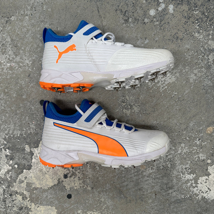 White Puma cricket bowling spikes with orange and blue accents on a gray surface.