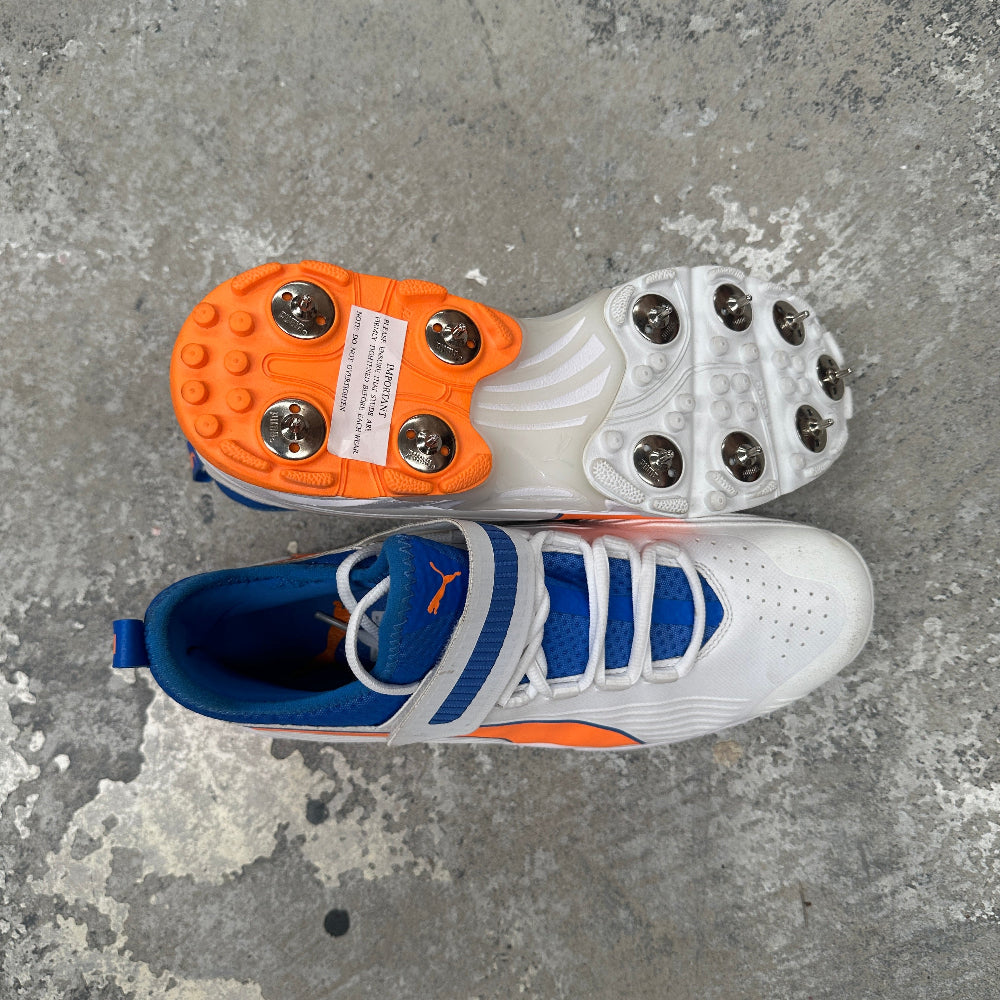Pair of blue and white Puma cricket spikes with orange accents on a gray concrete surface.